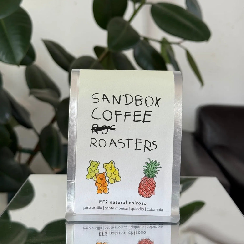 Sandbox Coffee Roasters packaging with coffee beans on a white surface with green leaves in the background