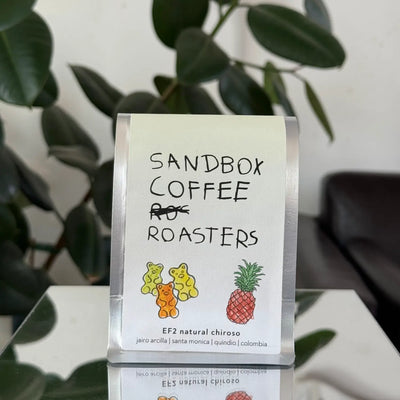 Sandbox Coffee Roasters packaging with coffee beans on a white surface with green leaves in the background