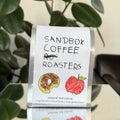 Sandbox Coffee Roasters coffee bag with illustrations of a donut and an apple on a stand.