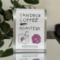Sandbox Coffee Roasters label with a purple flower graphic on a clear stand.