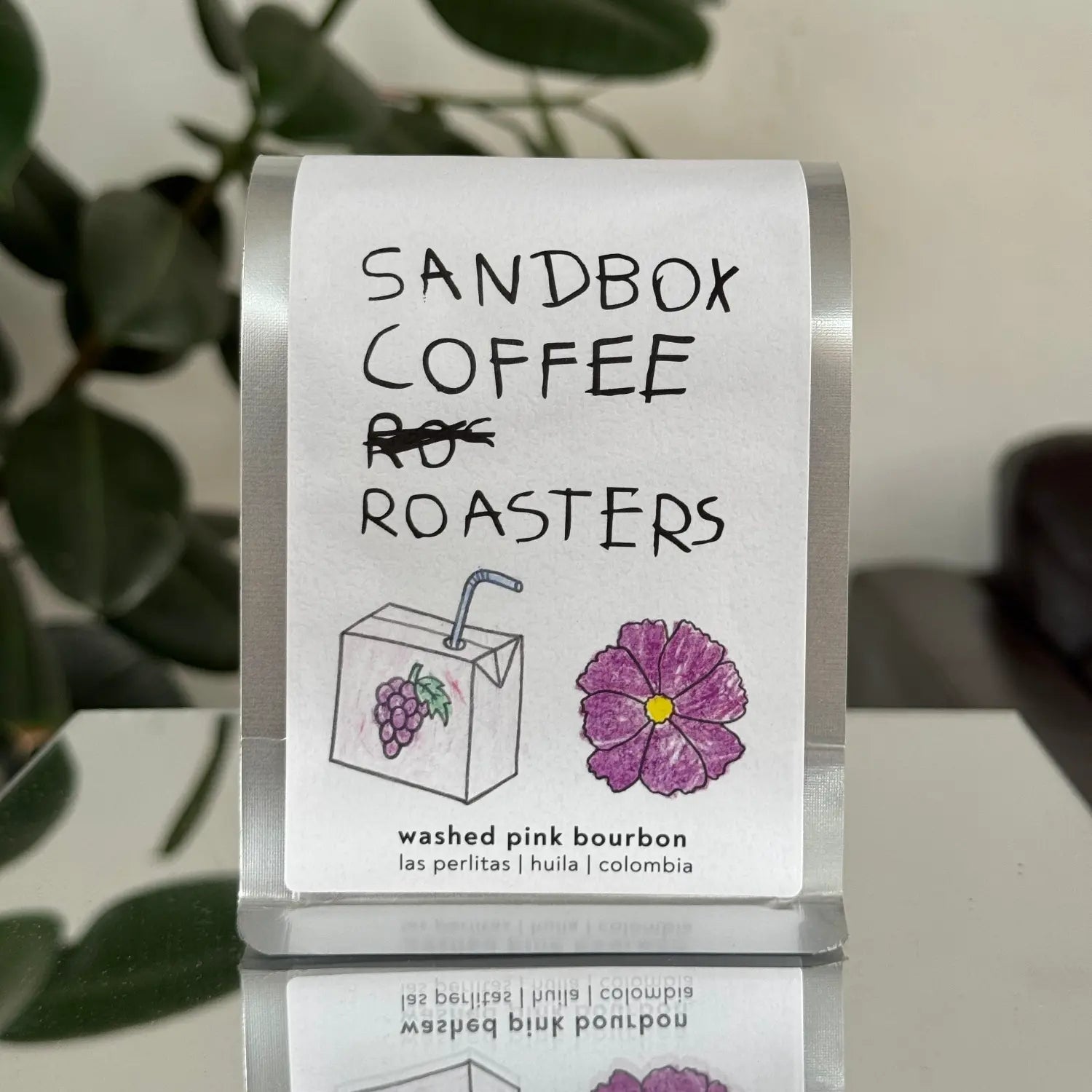 Sandbox Coffee Roasters label with a purple flower graphic on a clear stand.