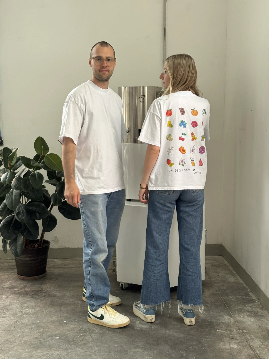 Two people in casual white t-shirts and jeans standing by a coffee roaster indoors