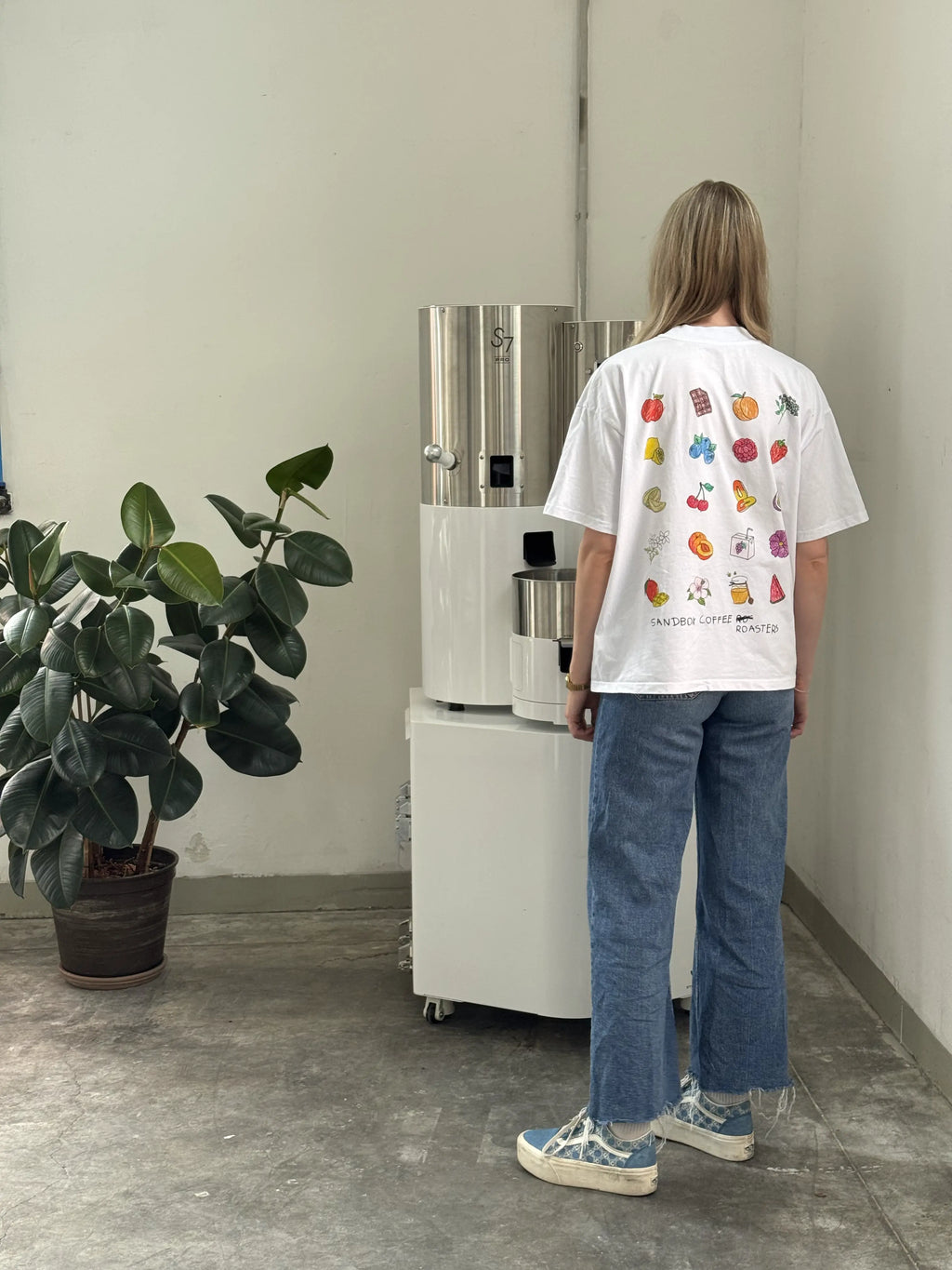 Person in a graphic white t-shirt and jeans standing by a modern coffee roaster in a minimal room.