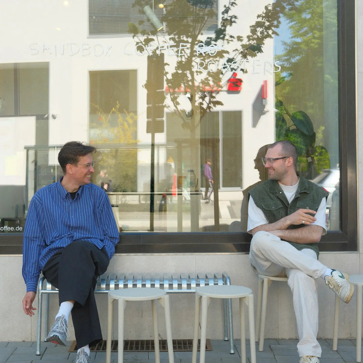 Two men sitting on stools outside a modern building with large glass windows.